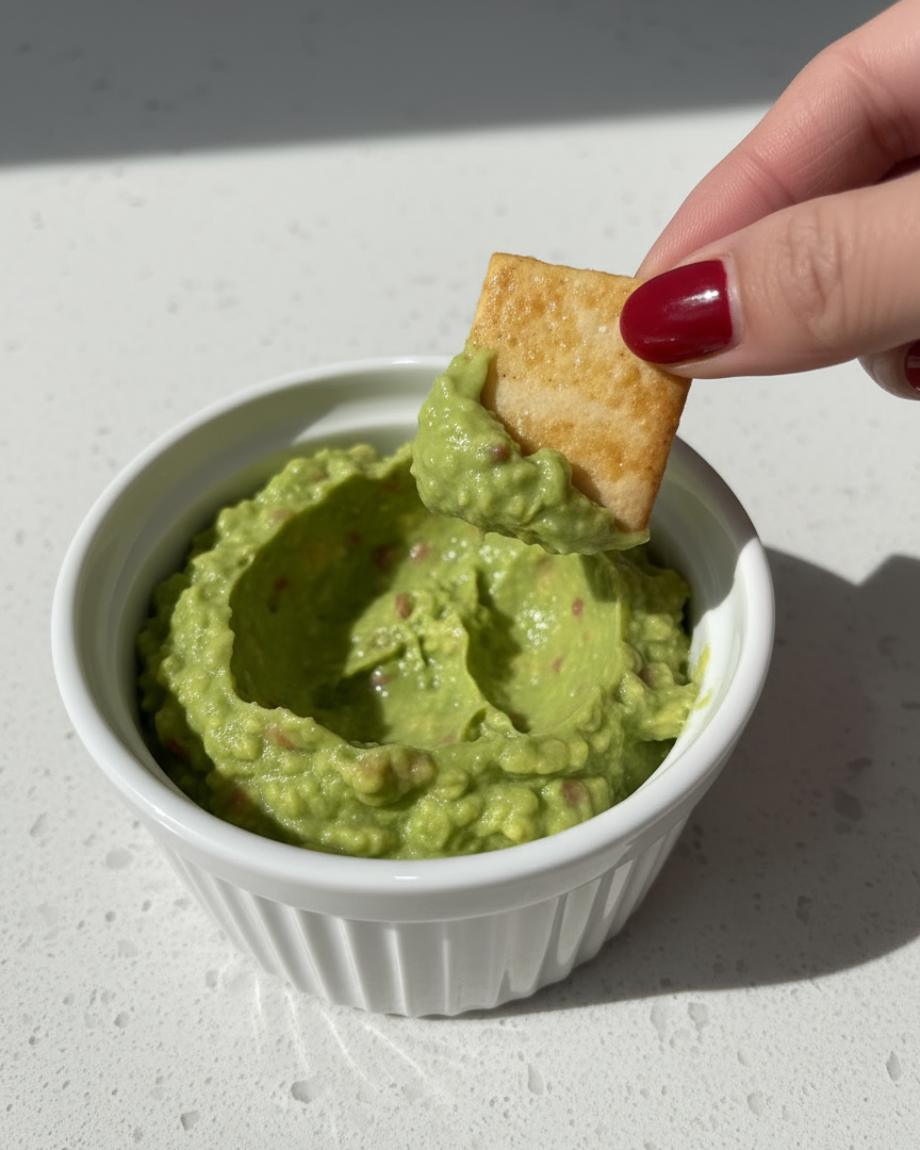 Hand dipping a chip into a bowl of guacamole on a light surface